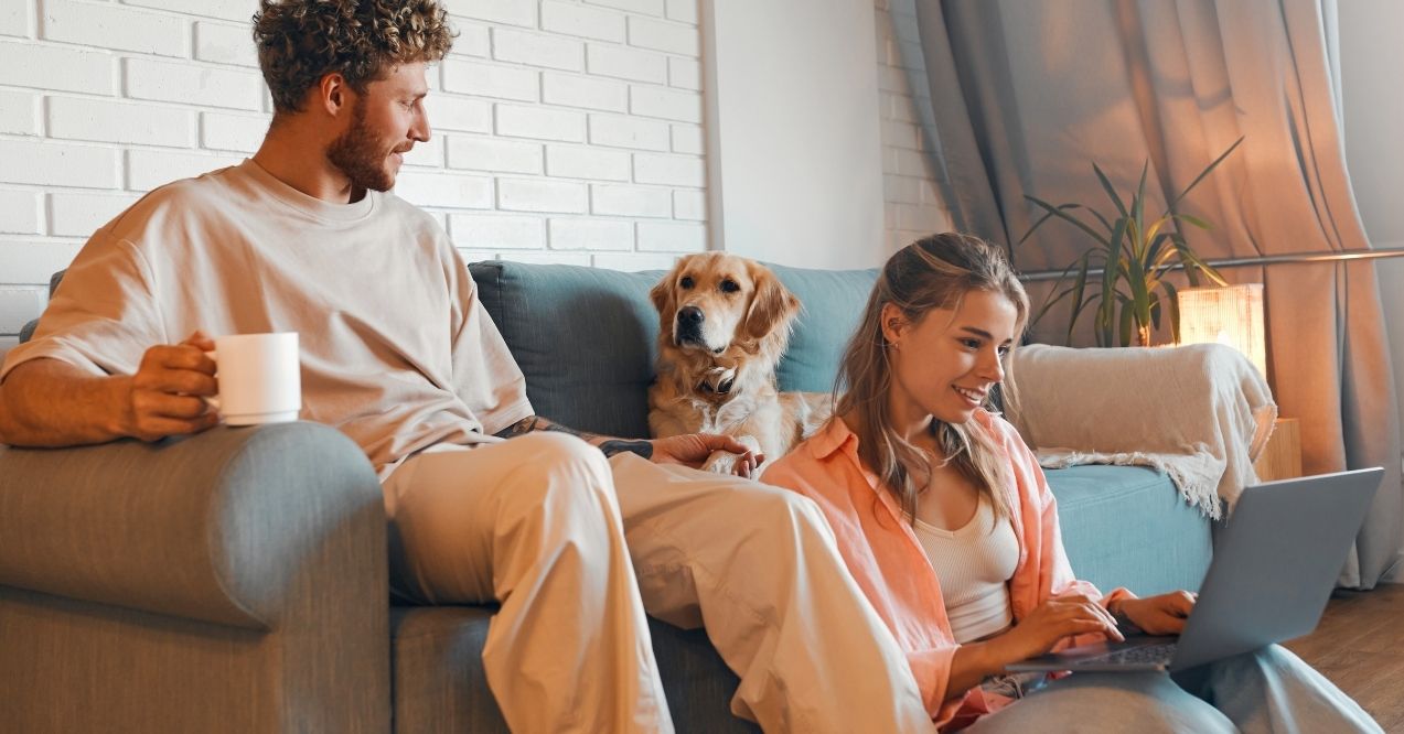 Man with coffee mug and woman on laptop with golden retriever on sofa